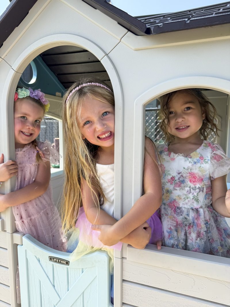 Three children smiling from the windows of the outdoor playhouse at Theodore's Academy